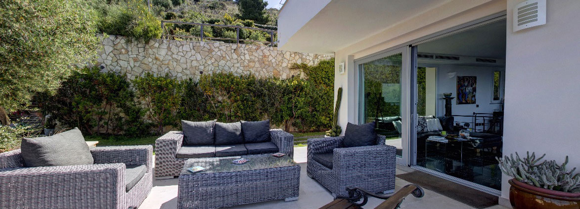 Outdoor seating area with wicker furniture and glass table, surrounded by greenery.