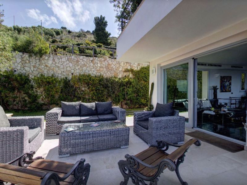 Outdoor seating area with wicker furniture and glass table, surrounded by greenery.