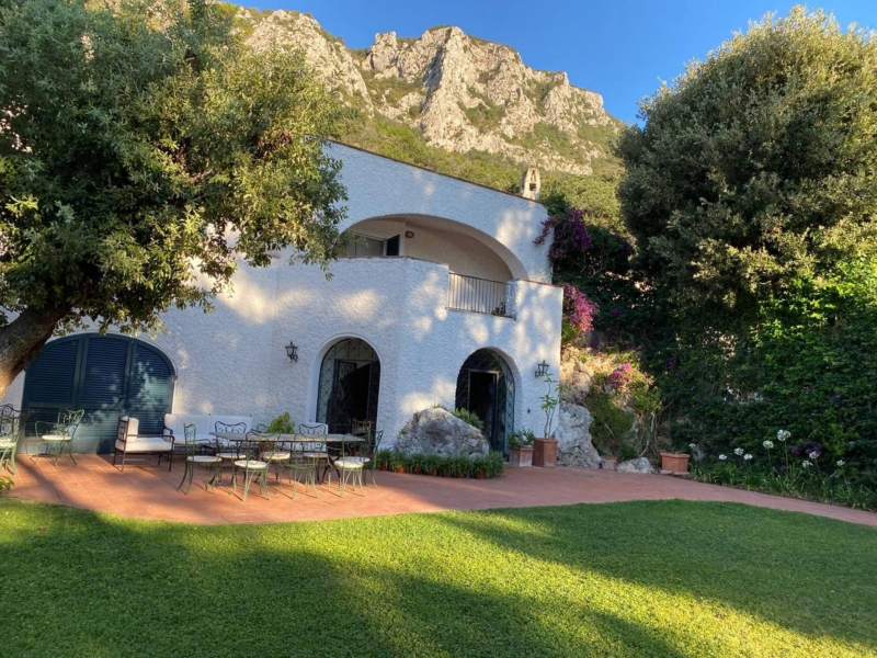 White villa with arched windows, outdoor seating, surrounded by greenery and mountains.