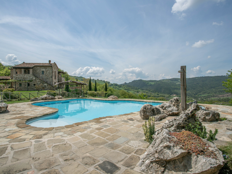 Stone house beside a blue pool, with lush green hills and clouds in the background.