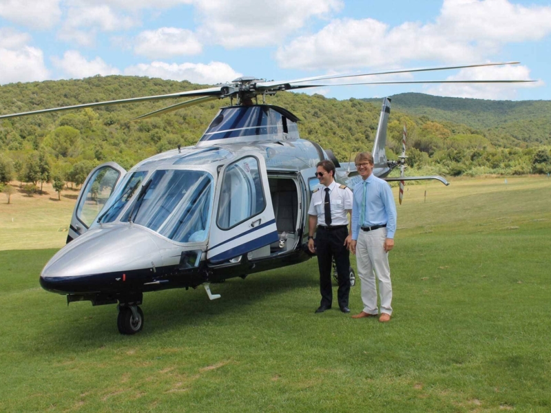 A pilot and a man stand beside a helicopter on a grassy field with hills in the background.