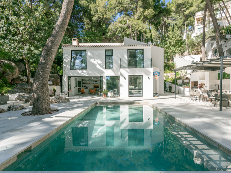 Modern white house with large windows, surrounded by greenery and a pool reflecting its structure.